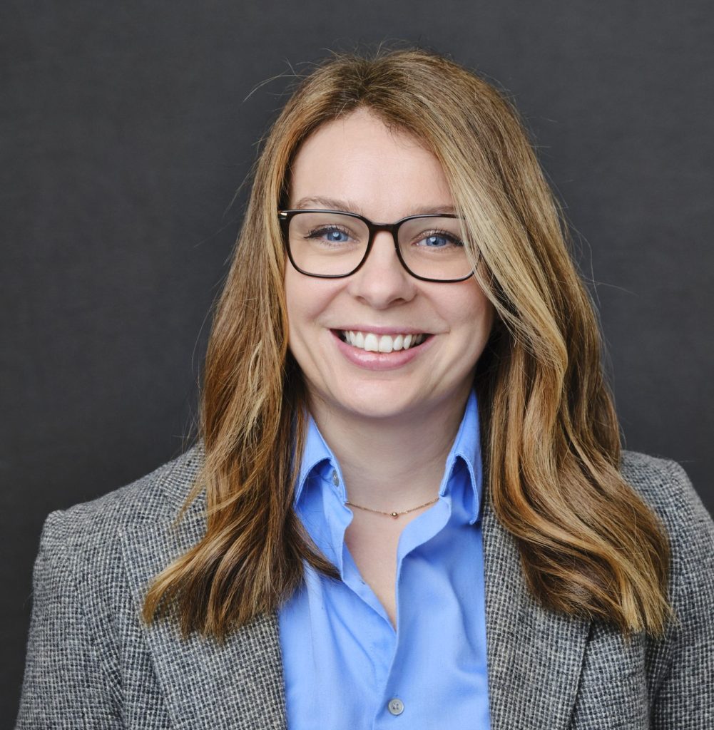 white person with blonde hair and glasses, blue shirt and grey blazer