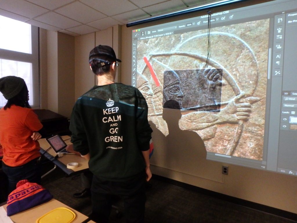 student with green short with text=keep calm and go green in front of projected image of egyptian archaeology on big screen